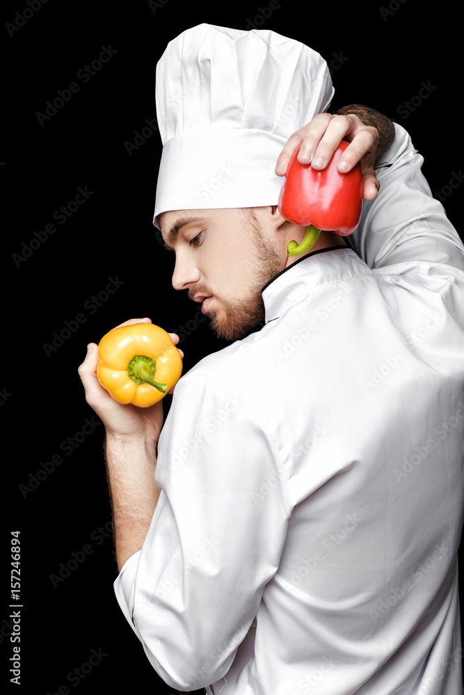 Young bearded man chef In white uniform holds bell peppers on black background Young bearded man chef In white uniform holds bell peppers on black background