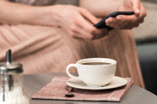 Close-up partial view of woman using smartphone and white cup with coffee on table