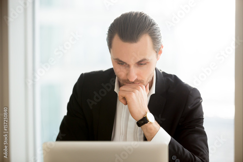 Serious businessman sitting in front of laptop and thinking about problem. Focused entrepreneur searching solution of difficult problem in internet. Man at computer ponders decision, working on report