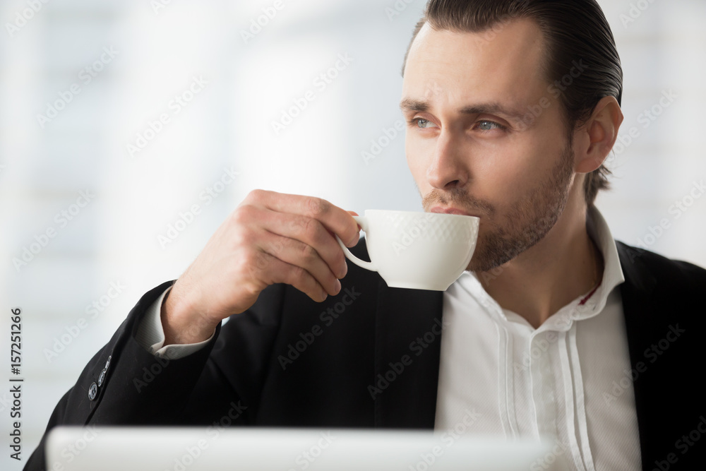 Portrait of young man drinking coffee and looking toward window. Businessman takes coffee-break at workplace. Thoughtful office worker holding white porcelain cup in hand dreaming about future success Portrait of young man drinking coffee and looking toward window. Businessman takes coffee-break at workplace. Thoughtful office worker holding white porcelain cup in hand dreaming about future success