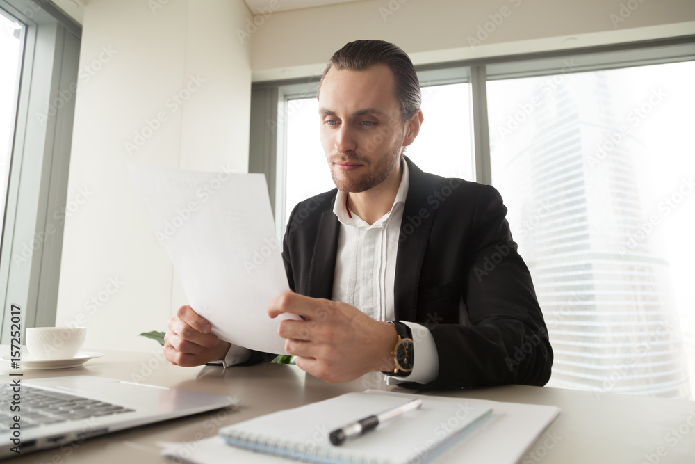 Successful businessman examines company financial documents at desk in office. Young entrepreneur reading contract at workplace. Executive manager analyzing report, resume, invoice, or business letter Successful businessman examines company financial documents at desk in office. Young entrepreneur reading contract at workplace. Executive manager analyzing report, resume, invoice, or business letter