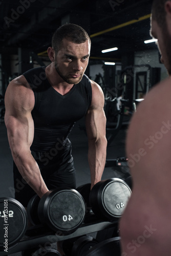 young sportsman holding dumbbells while looking at reflection in mirror