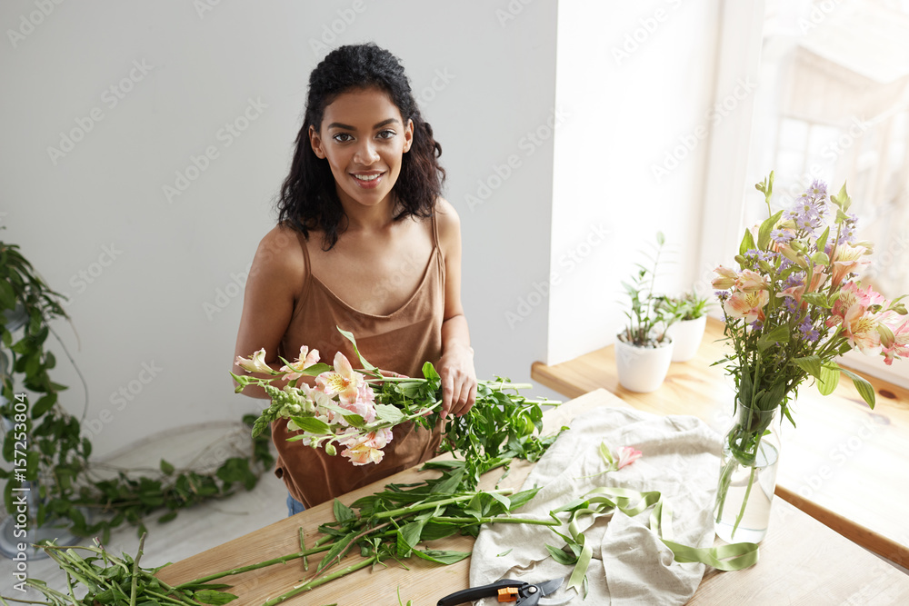 Young beautiful african girl florist taking care of flowers at workplace over white wall. Young beautiful african girl florist taking care of flowers at workplace over white wall.