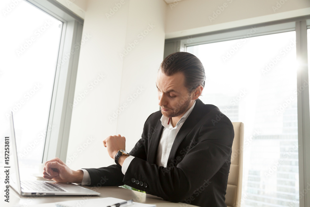 Office worker at desk looking at wristwatch, waiting for the end of work day. Busy young entrepreneur hurrying to finish computer report. Businessman checks time remaining to important office meeting Office worker at desk looking at wristwatch, waiting for the end of work day. Busy young entrepreneur hurrying to finish computer report. Businessman checks time remaining to important office meeting