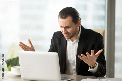Billede på lærred Upset shocked businessman at office table looks at laptop screen with surprised confused expression, lost finance data