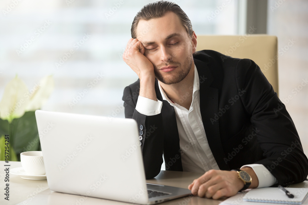 Young man dozing with head on hand while sitting at desk with laptop in office. Businessman sleeping at workplace in morning after weekend party day before. Tired male entrepreneur slumbers at work Young man dozing with head on hand while sitting at desk with laptop in office. Businessman sleeping at workplace in morning after weekend party day before. Tired male entrepreneur slumbers at work