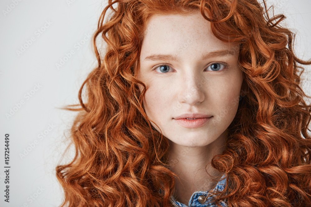 Close up of beautiful girl with curly red hair and freckles looking at camera over white background.