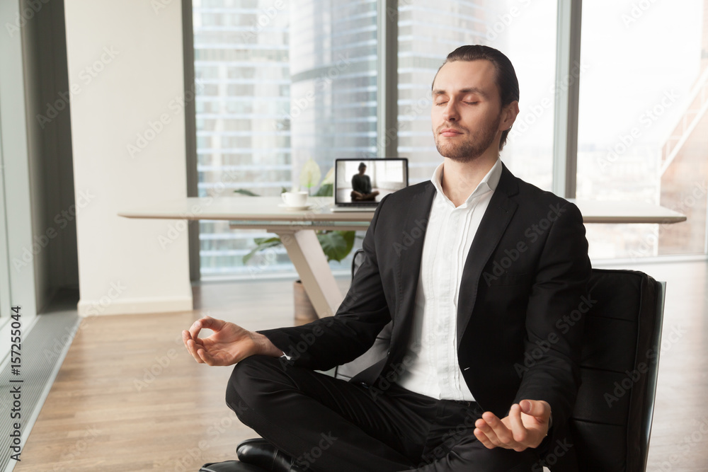 Businessman meditates in lotus position on coach in modern office. Company leader practicing yoga during break at work for strength recovery and stress relief. Man relaxing near workplace with laptop Businessman meditates in lotus position on coach in modern office. Company leader practicing yoga during break at work for strength recovery and stress relief. Man relaxing near workplace with laptop