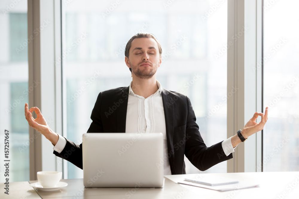 Relaxed young entrepreneur meditating at desk in front of laptop. Office worker doing yoga exercises at workplace. Successful businessman relieves stress with spiritual practices or autosuggestion Relaxed young entrepreneur meditating at desk in front of laptop. Office worker doing yoga exercises at workplace. Successful businessman relieves stress with spiritual practices or autosuggestion