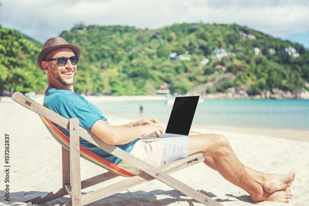 Attractive young man with laptop working on the beach. Freedom, remote work, freelancer, technology, internet, travel and vacation concepts