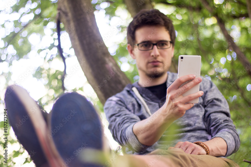 Junger Mann lehnt im Park am Baum, Blick aufs Smartphone Junger Mann lehnt im Park am Baum, Blick aufs Smartphone
