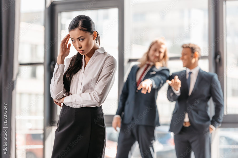 frustrated asian businesswoman standing in office, businessmen behind gesturing and laughing frustrated asian businesswoman standing in office, businessmen behind gesturing and laughing