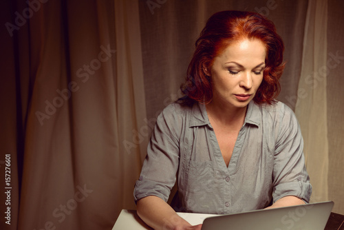 pensive red hair woman working on laptop while sitting at table at home