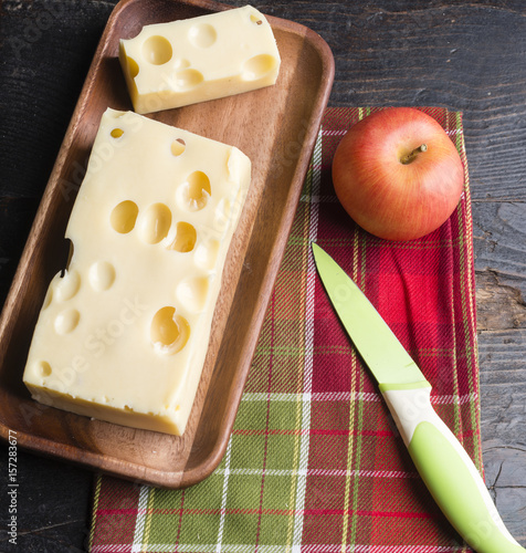 A piece of emmethal swiss cheese over a wooden rustic plate, a green knife and a fresh appe.