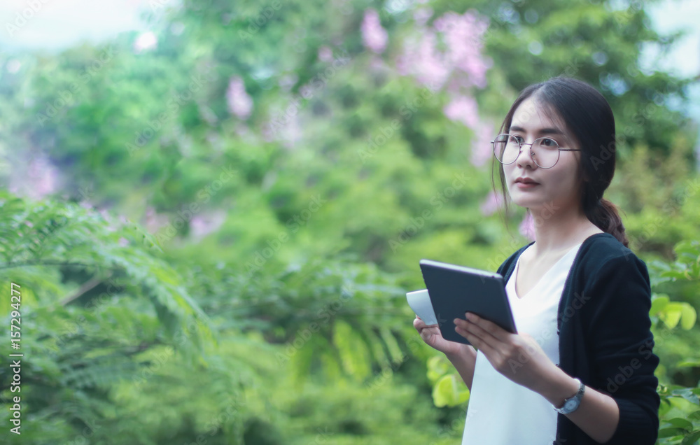 Fototapeta premium Happy young student with a tablet and a disposable coffee cup sitting on the bench and reading in a summer park.