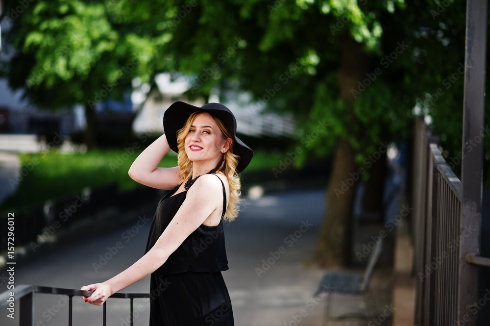 Blonde woman on black dress, necklaces and hat posed at streets.