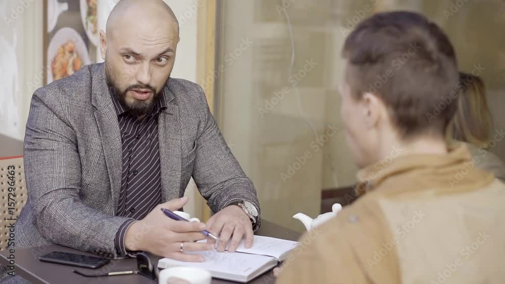 Business partners discuss the development strategy of making a note in my diary on a sheet of paper with a ballpoint pen. Opponent with a bald head and a beard says gesturing emotionally. Business partners discuss the development strategy of making a note in my diary on a sheet of paper with a ballpoint pen. Opponent with a bald head and a beard says gesturing emotionally.