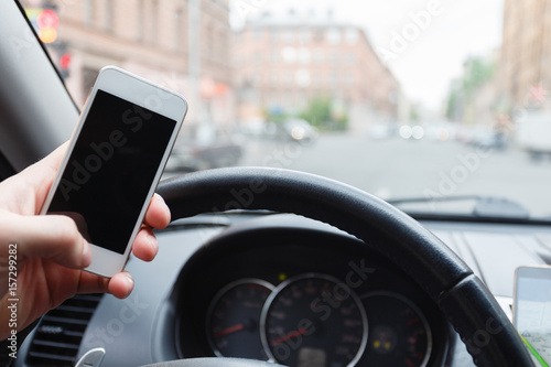 Hand with mobile phone on the steering wheel. Using a smartphone while driving. Is in motion in city traffic. © korchemkin