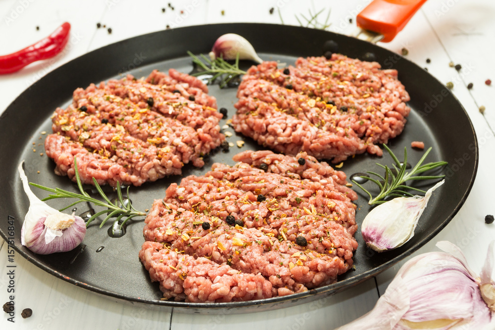 Organic beef raw cutlets with spices in a frying pan on a white background with garlic, rosemary and pepper.