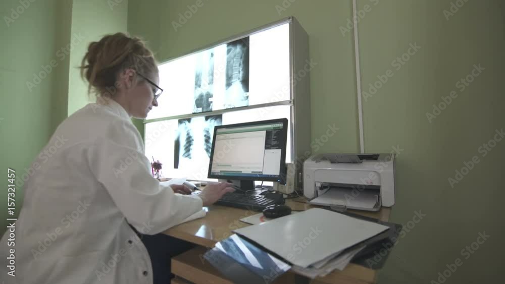Female doctor specialist, radiologist in white gown is sitting at a computer and typing on the keyboard medical reports during examining x-rays on the light board, steady cam shot, wide angle lens. Female doctor specialist, radiologist in white gown is sitting at a computer and typing on the keyboard medical reports during examining x-rays on the light board, steady cam shot, wide angle lens.
