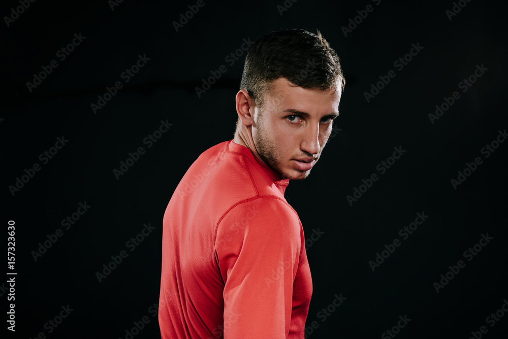 Profile view of muscular athlete man in red sportswear standing over dark background, thinking over problems in his life, holding hands on his waist, looking away. Lifestyle, sport, motivation.