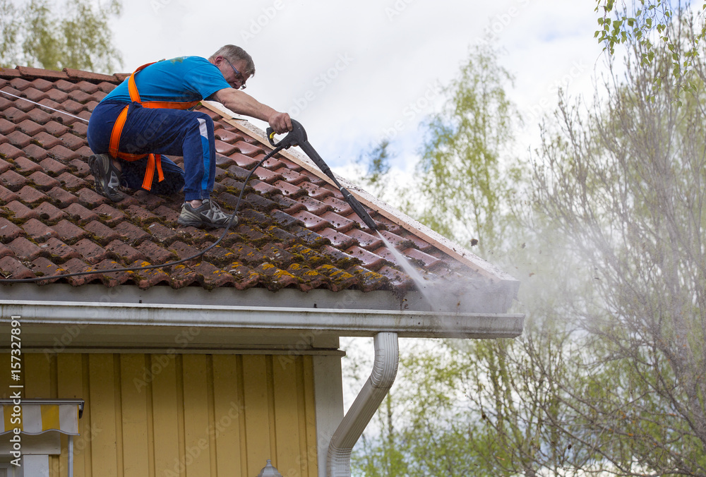 Caucasian man is washing the roof with a high pressure washer. He is  wearing safety harness on a slippery roof. Stock Photo