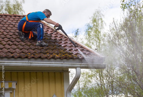Caucasian man is washing the roof with a high pressure washer. He is wearing safety harness on a slippery roof. photo