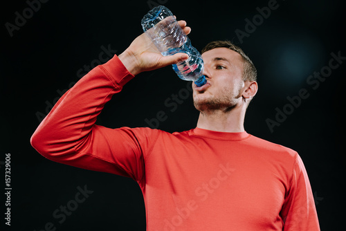 Attractive young Caucasian sportsman drinking water from bottle looking far away with thoughtful face expression, dressed in red sportswear, relaxing after run or workout. Sport motivation. Lifestyle.