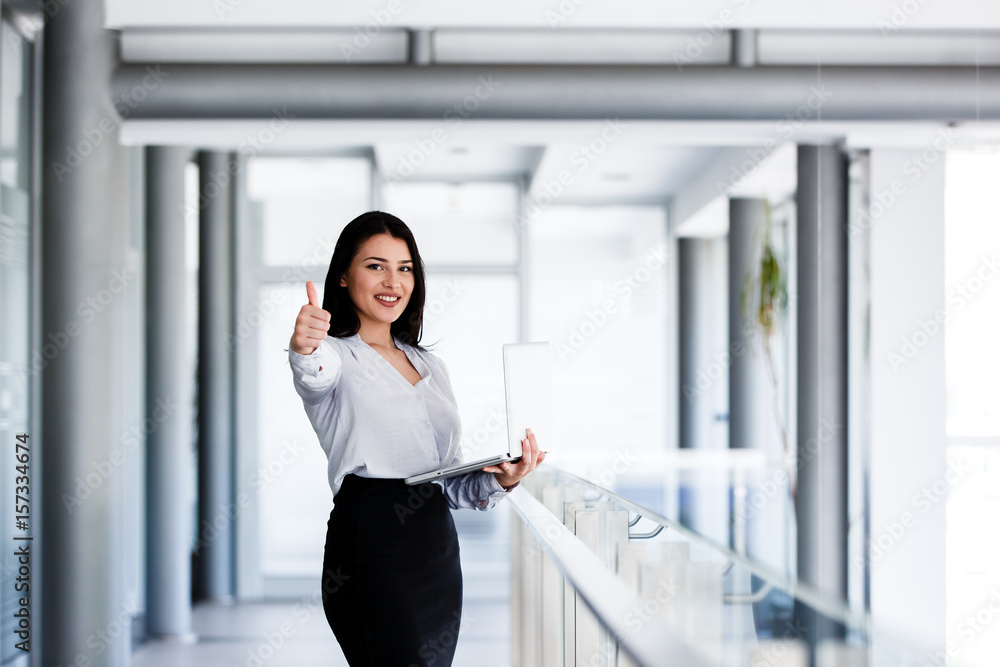 Attractive and smiling woman showing thumbs up while holding laptop in her hand Attractive and smiling woman showing thumbs up while holding laptop in her hand