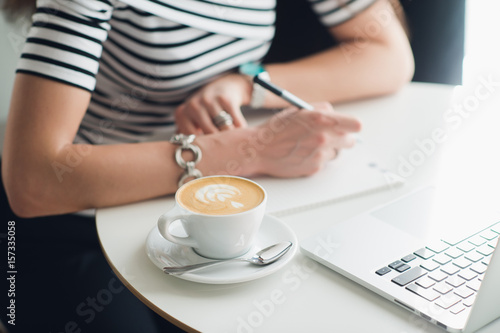 A white blank notebook with a luxury pen and a cup of Latte coffee with blurred laptop in the background.