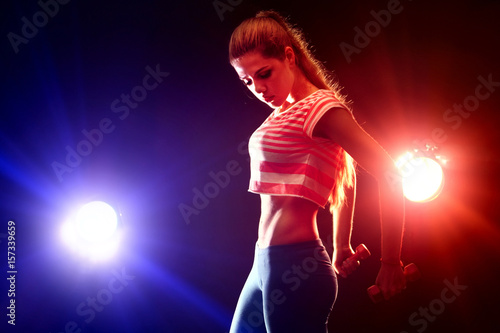 Fitness girl exercising in gym with dumbbells. Woman has slim stomach works in multi-colored rays of sport room. Portrait of full-length in profile. Flat stomach as motivation for sports