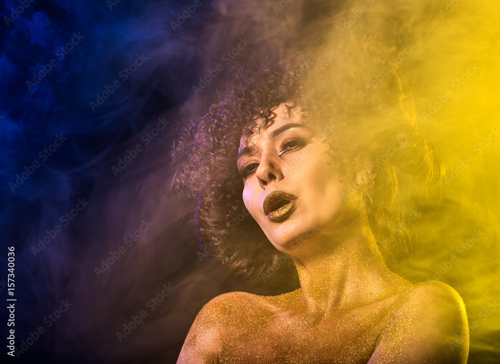 Golden powder cosmetics on bare woman shoulders with decorative. Girl with curls on dark background. Woman at disco in gold mystical fog . Smoke from hookah.