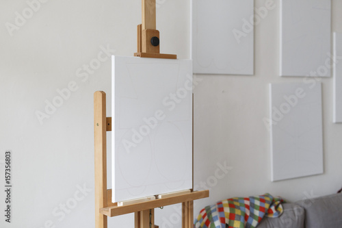 an empty canvas on an easel with multiple empty canvasses on the wall behind photo