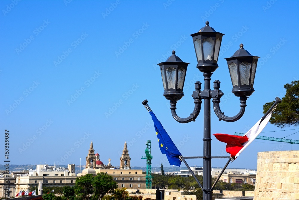 Maltese and EU flag on a lamppost with city buildings to the rear, Valletta, Malta.
