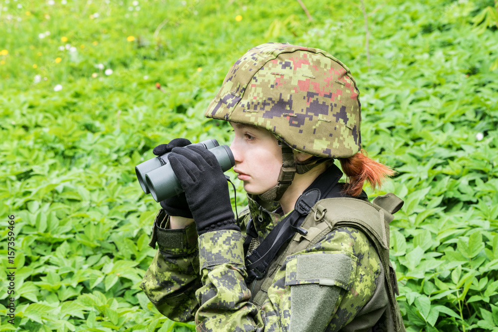 Portrait of armed woman with camouflage. Young female soldier observe with binoculars. Child soldier with gun in war, green goutweed background.  Military, army people concept