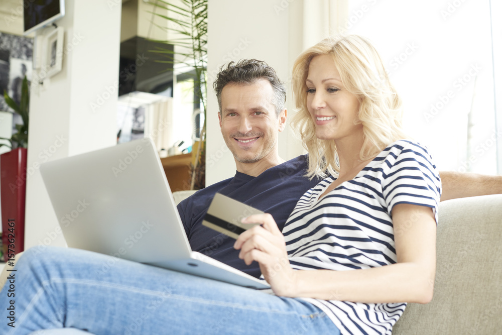 They like to shop online. Shot of a smiling middle aged couple sitting on couch and using credit card and laptop while shopping online. They like to shop online. Shot of a smiling middle aged couple sitting on couch and using credit card and laptop while shopping online.
