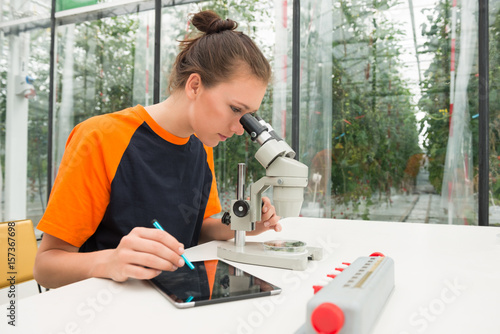 Young female botanist examining samples of plant under microscope while using digital tablet at table