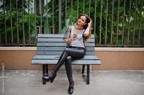 Fashionable woman look at white shirt, black transparent clothes, leather pants, posing at street and sitting on bench with cell phone and headphones. fashion girl listening music on mobile.