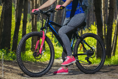 One young woman - an athlete in the pink shoes walks with the mountain bike in a pine forest in the morning .