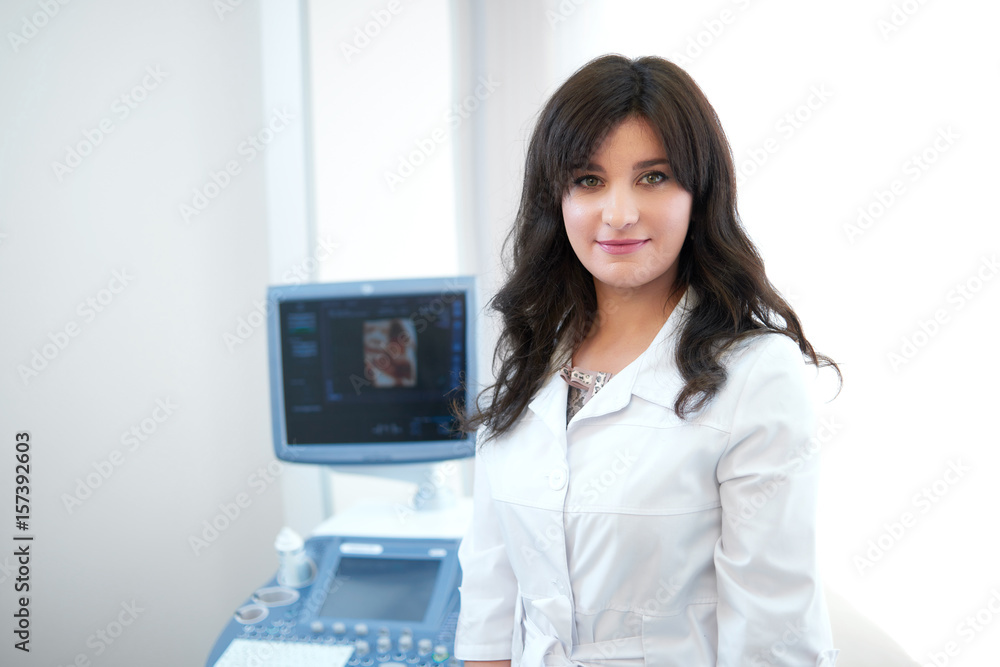 Pretty brunette female doctor standing against ultrasound equipment and computer, looking, smiling at camera. Professional in white uniform posing in cabinet at modern hospital. Medicine concept. Pretty brunette female doctor standing against ultrasound equipment and computer, looking, smiling at camera. Professional in white uniform posing in cabinet at modern hospital. Medicine concept.