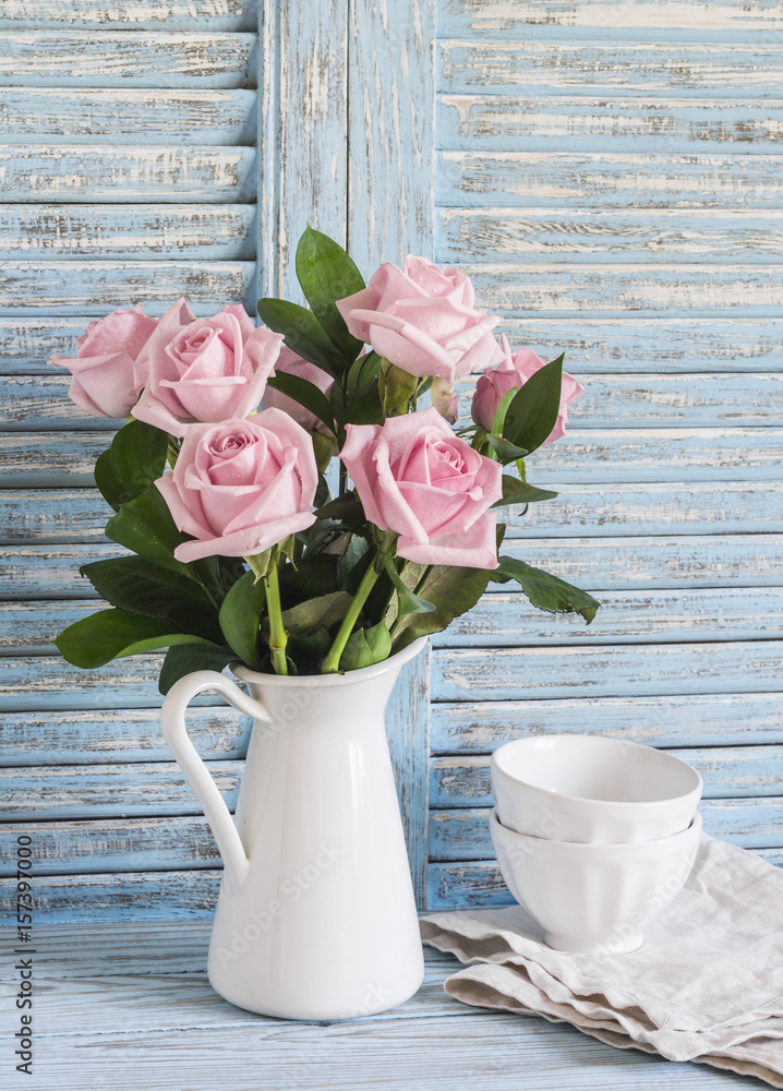 Fototapeta premium Pink roses in a white enameled pitcher and ceramic white bowls on blue wooden rustic background. Kitchen still life in vintage style