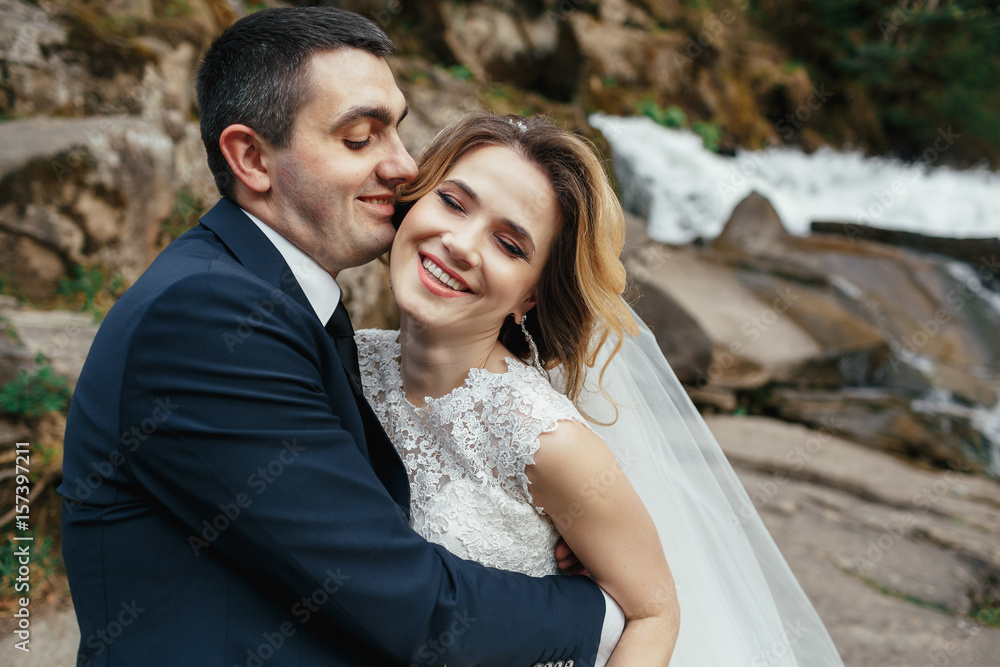 Hugging wedding couple stands before a waterfall