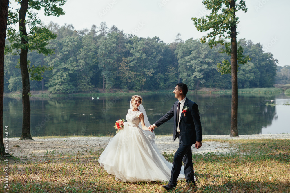 Groom holds bride's hand walking along the shore of a lake