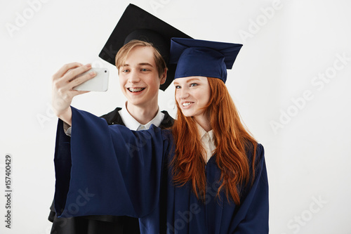 Friends graduates of college in caps smiling making selfie over white background.