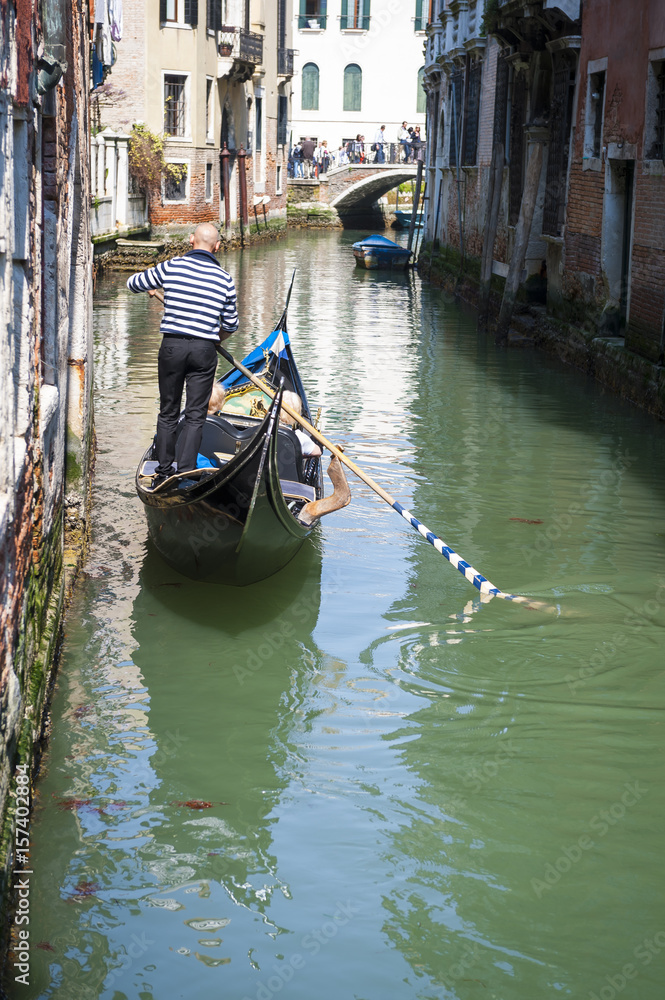 Scenic view of a small canal in Venice, Italy, with gondolier in traditional striped shirt maneuvering gondola along green waters