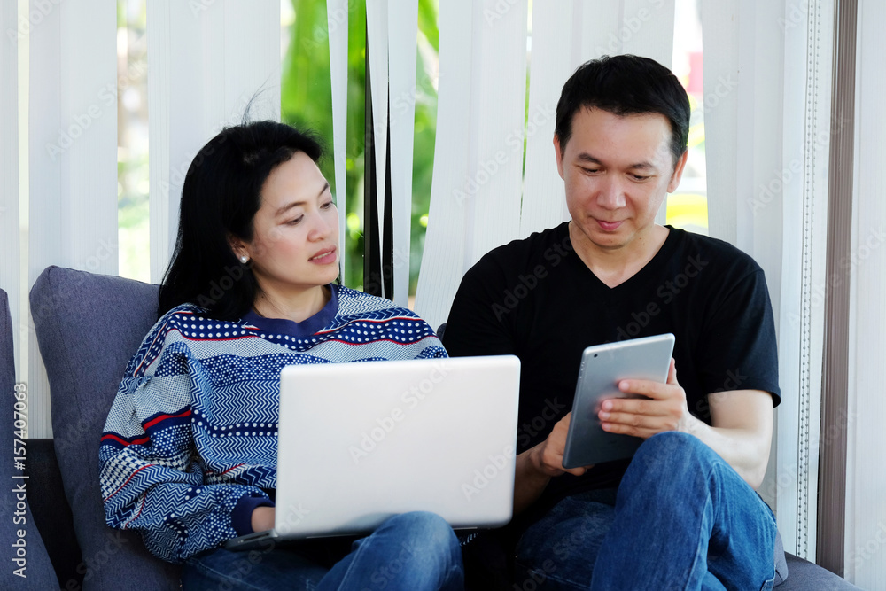 The couple using lab-top computer and tablet sitting on sofa at home, lifestyle, internet of things concept The couple using lab-top computer and tablet sitting on sofa at home, lifestyle, internet of things concept