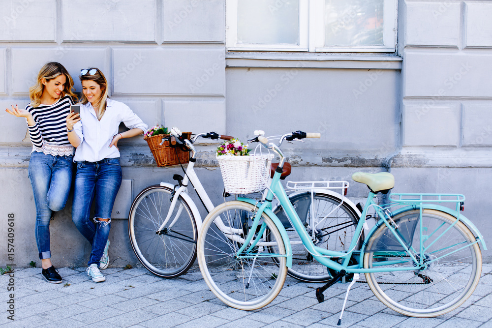 Fototapeta premium Two young women with bikes using smartphone