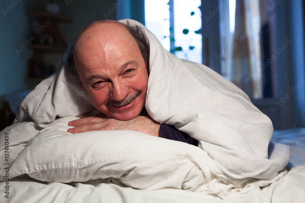 An elderly bald man with a mustache lies in bed, hugging a pillow. He has a problem with sleep. An elderly bald man with a mustache lies in bed, hugging a pillow. He has a problem with sleep.