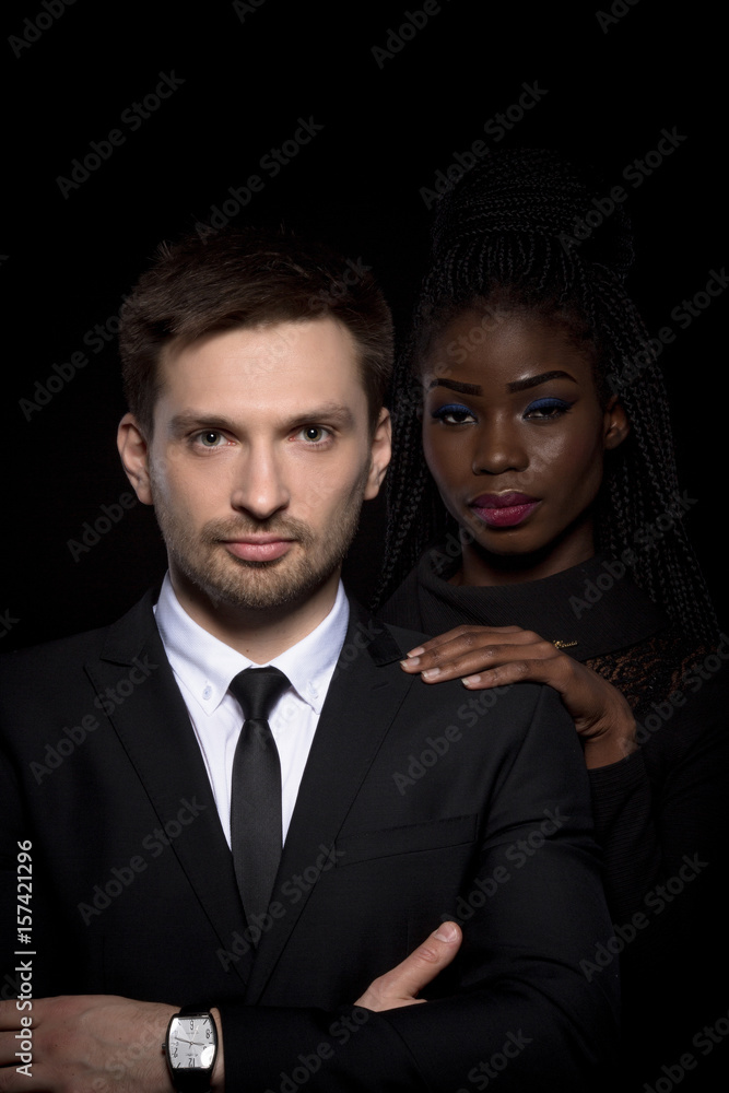 Close up portrait of multi-ethnic couple on black background. Multi-ethnic man and woman posing on black backstage. Man in business suit with dark skinned woman behind.