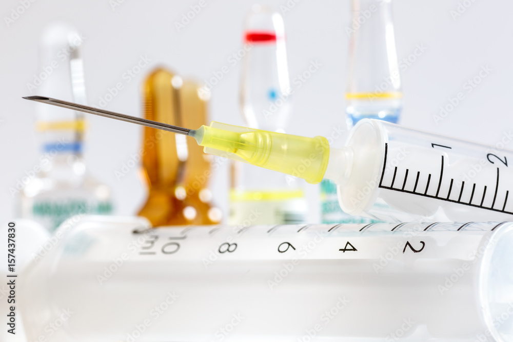 Close-up of needle of hypodermic syringe in front of ampules of drugs, on white Close-up of needle of hypodermic syringe in front of ampules of drugs, on white